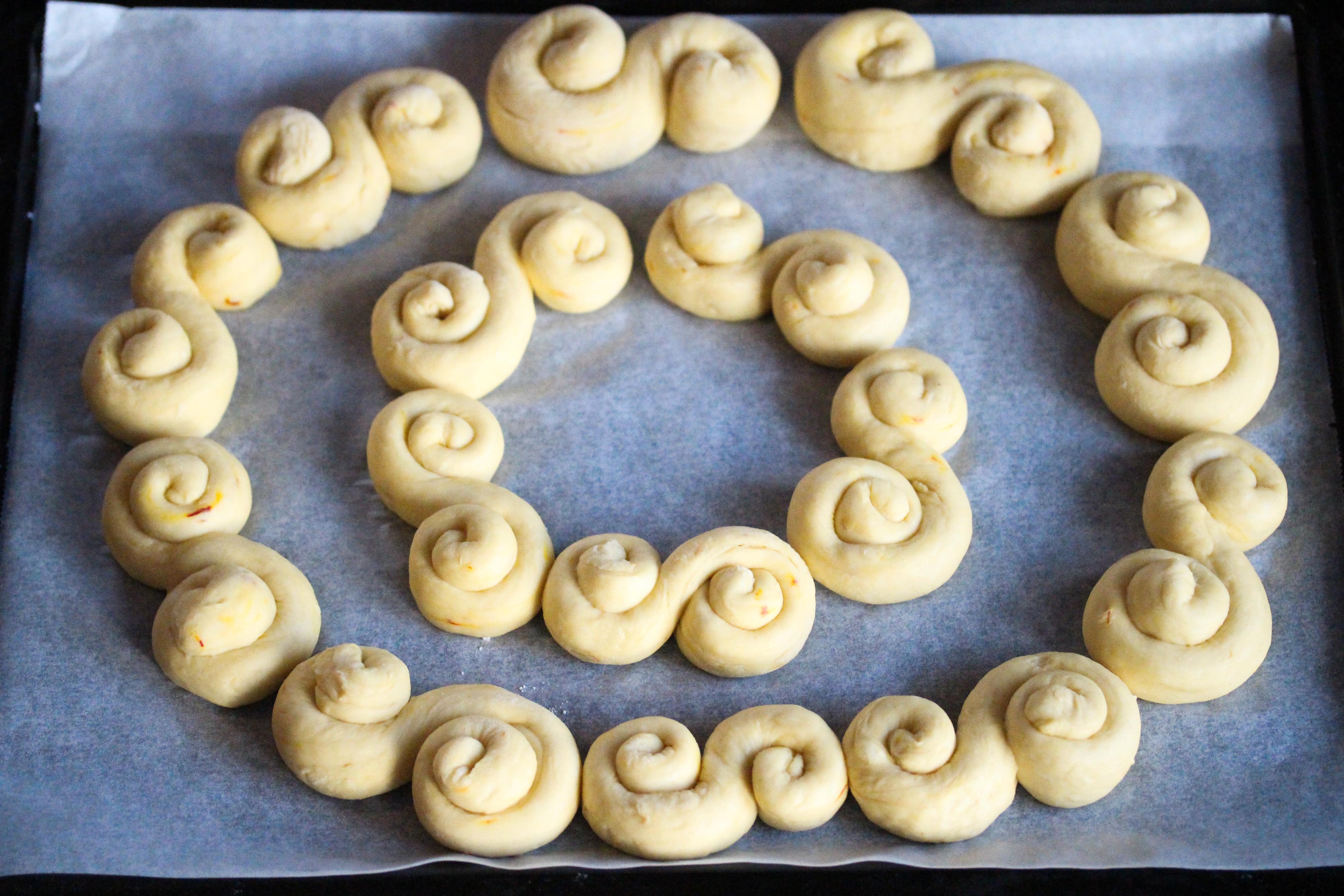 Del deigen i mindre emner, som trilles til lengder og som formes til S’er. Dekk en stekeplate med bakepapir og legg lussekattene i en stor krans og deretter i en indre krans. Jeg brukte 10 lussekatter i den ytre kransen og 5 i den indre, men dette avhenger av hvor store du lager hver Lussekatt. Husk at Lussekattene hever seg en god del under stekingen, så la det være litt luft mellom dem. Etterhev kransen i 30 minutter.