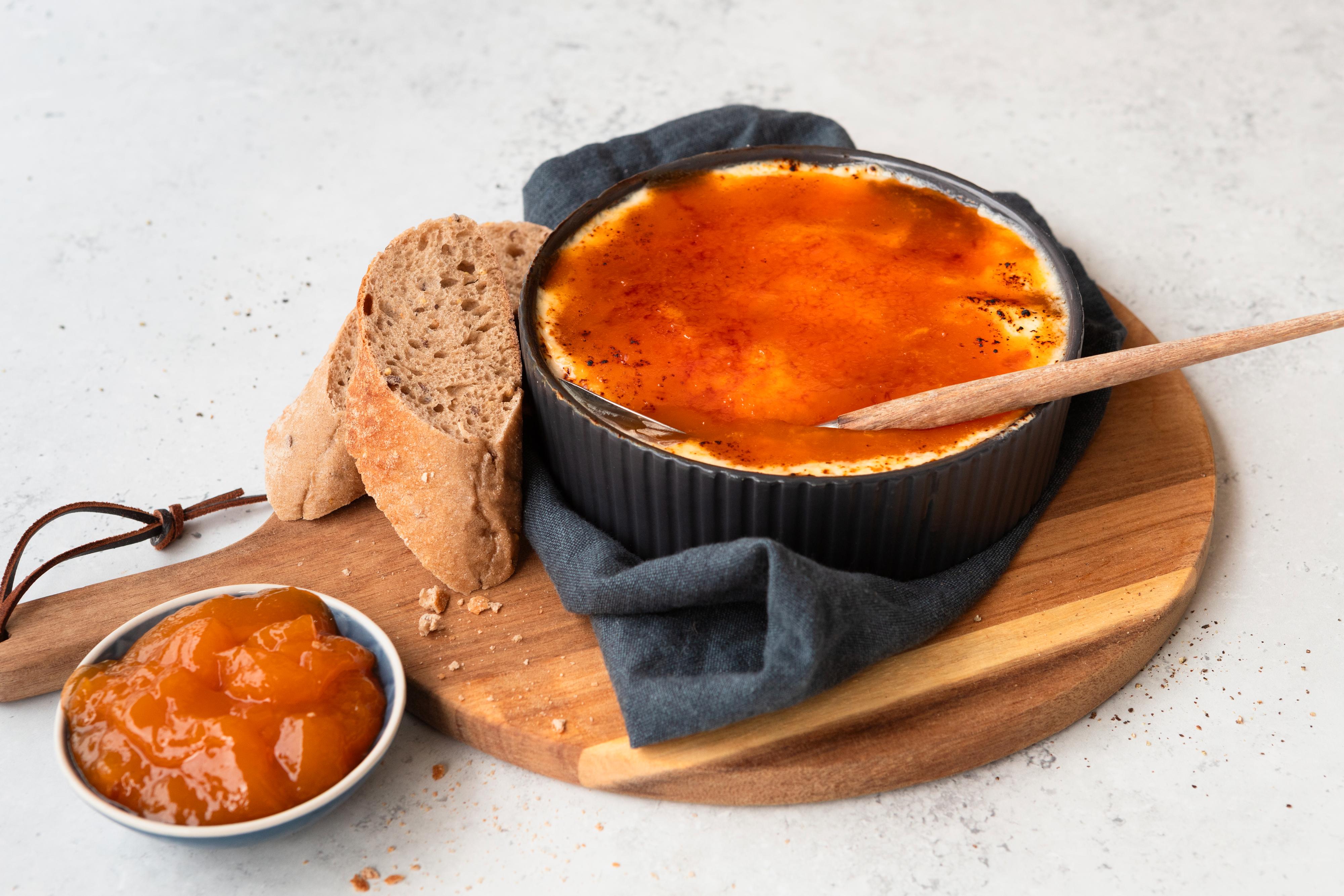 På en rundt trefjøl står en liten, svart ildfast form med ovnsbakt ost og aprikosslokk. Ved siden av er noen skiver baguett og en liten skål med aprikossyltetøy.