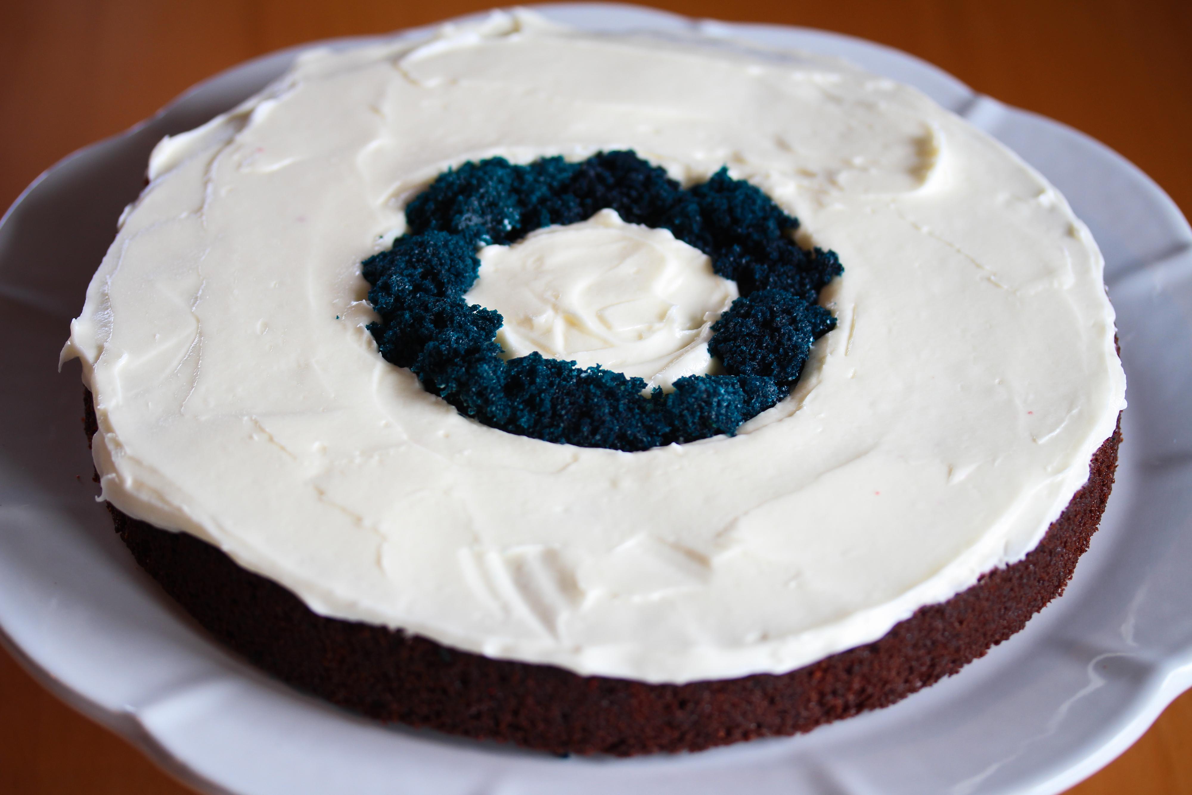 Smør ostekrem over de røde kakeflatene. Pass på så det ikke kommer noe ostekrem på blå kake. Fyll opp mellomrommet mellom den blå kakesirkelen og ostekrem med litt blå kakesmuler, så den blå kakesirkelen får samme høyde som resten av kaken.